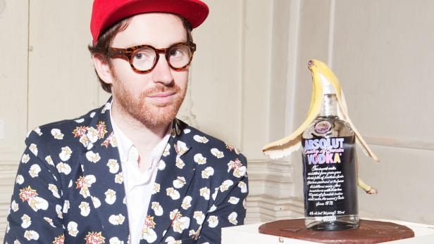 Ein Mann mit roter Kappe und Popcorn-Jacke posiert neben einer Absolut Vodka Flasche mit Bananenschale.