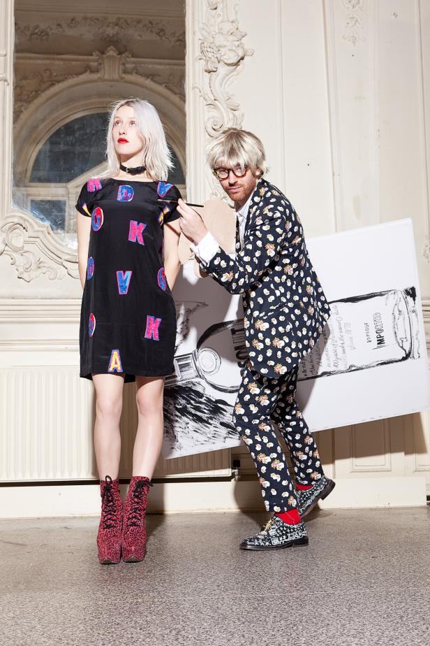 Eine Frau in einem Kleid mit Buchstaben posiert, während ein Mann mit Perücke ein großes Bild hält.