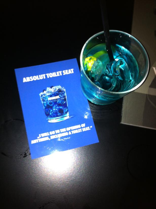 Ein blaues Getränk mit Eiswürfeln neben einer „Absolut Toilet Seat“-Werbung.