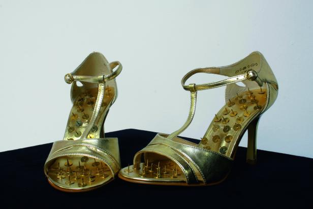 Ein Paar goldene High Heels mit Nägeln auf der Innensohle stehen auf einem schwarzen Tuch.