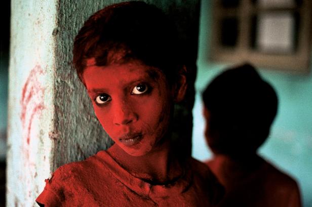 Ein Junge, dessen Gesicht und Kleidung mit roter Farbe bedeckt sind, lehnt an einer Wand.