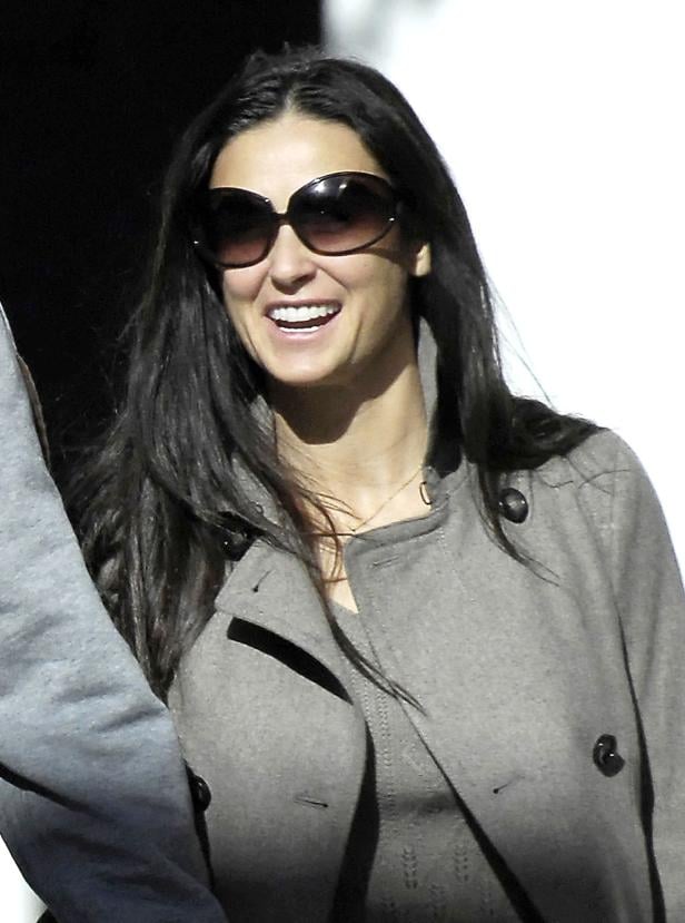 Demi Moore lächelt und trägt eine Sonnenbrille und einen grauen Mantel.