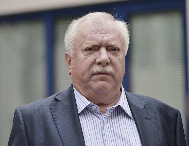 Porträt eines Mannes mit grauem Haar und Schnurrbart in einem blauen Anzug.