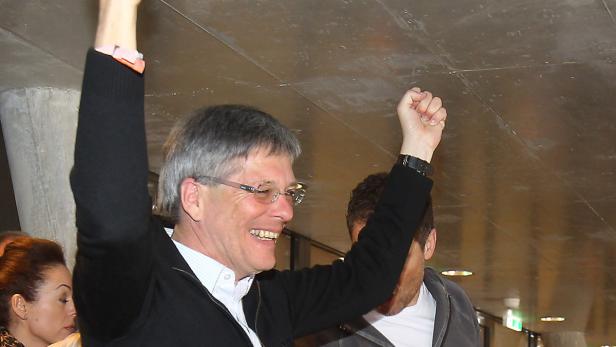Ein Mann mit Brille und schwarzer Jacke jubelt mit erhobenen Armen.