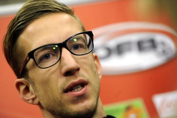Ein Mann mit Brille vor einem roten Hintergrund mit einem Logo.
