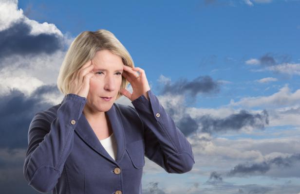 Eine Frau mit blonden Haaren hält sich die Schläfen vor einem Himmel mit Wolken.