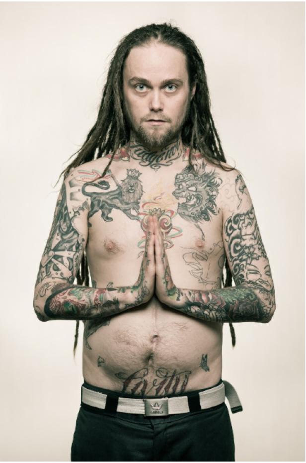 Ein tätowierter Mann mit Dreadlocks hält die Hände vor der Brust gefaltet.