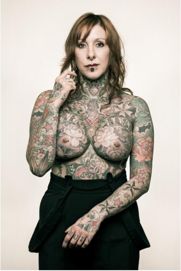 Eine tätowierte Frau mit Piercings posiert vor einem hellen Hintergrund.