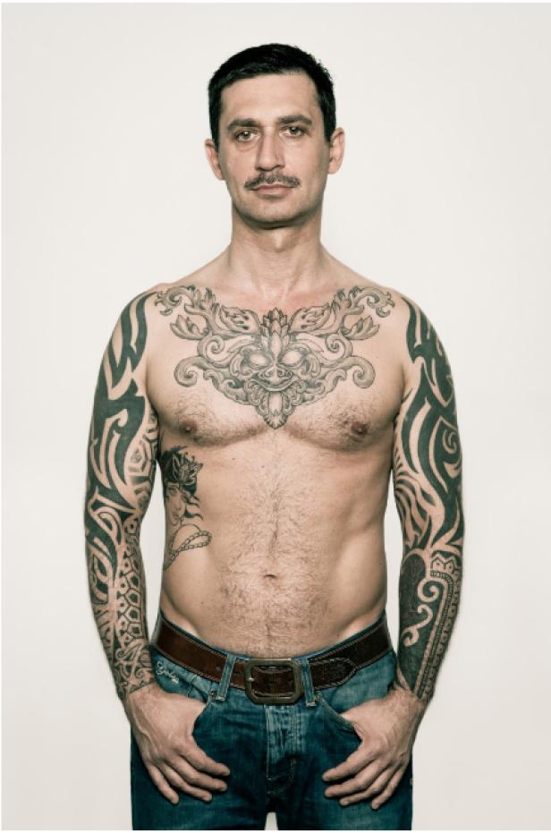 Ein Mann mit Tätowierungen am Oberkörper und an den Armen steht vor einer weißen Wand.