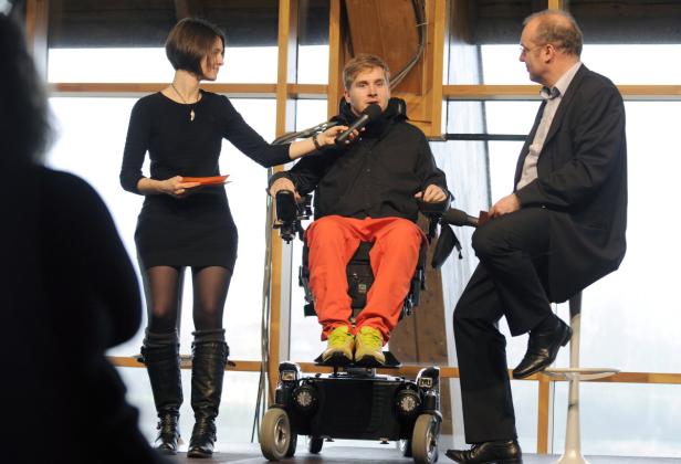 Ein Mann im Rollstuhl spricht in ein Mikrofon, während eine Frau ihm zuhört und ein Mann auf einem Barhocker sitzt.