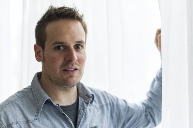 Ein Mann in einem hellblauen Hemd steht an einem Fenster mit weißen Vorhängen.