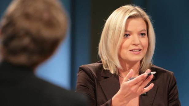 Eine blonde Frau in einem braunen Blazer gestikuliert mit einem Stift.