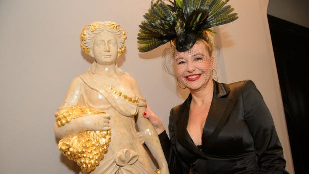 Eine Frau mit einem extravaganten Hut posiert neben einer Statue mit goldenen Akzenten.