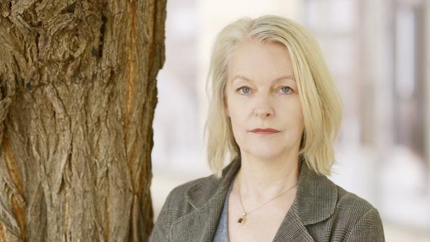 Eine Frau mit blonden Haaren steht neben einem Baumstamm.