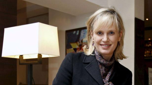 Eine blonde Frau lächelt in die Kamera, gekleidet in einen schwarzen Mantel und einen gemusterten Schal.