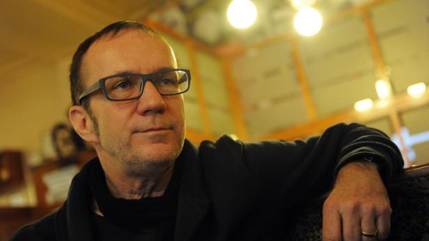 Ein Mann mit Brille und dunkler Jacke sitzt in einem Raum mit warmem Licht.