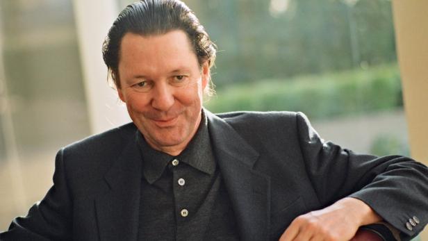 Ein lächelnder Mann in einem dunklen Anzug sitzt in einem Sessel.