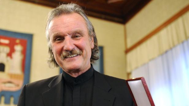 Ein lächelnder Mann mit Schnurrbart und dunklem Anzug hält eine Auszeichnung.