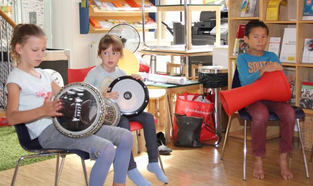 Drei Kinder sitzen auf Stühlen und spielen auf Trommeln.