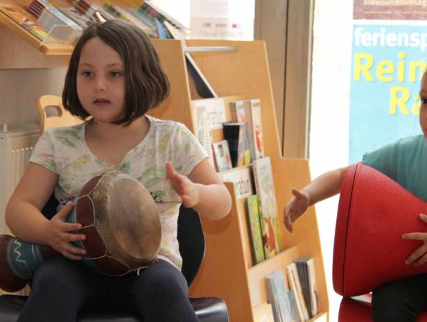 Ein Mädchen spielt auf einer Trommel, während ein anderes Kind daneben sitzt.