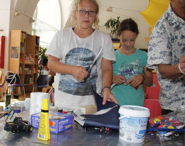 Zwei Mädchen basteln an einem Tisch mit verschiedenen Materialien wie Stoff und Klebstoff.