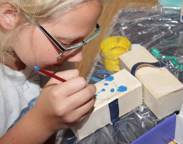 Ein Mädchen bemalt mit einem Pinsel einen Holzschuh mit blauer Farbe.