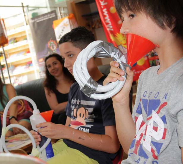 Zwei Jungen experimentieren mit Trichtern und Schläuchen, während eine Frau im Hintergrund zusieht.