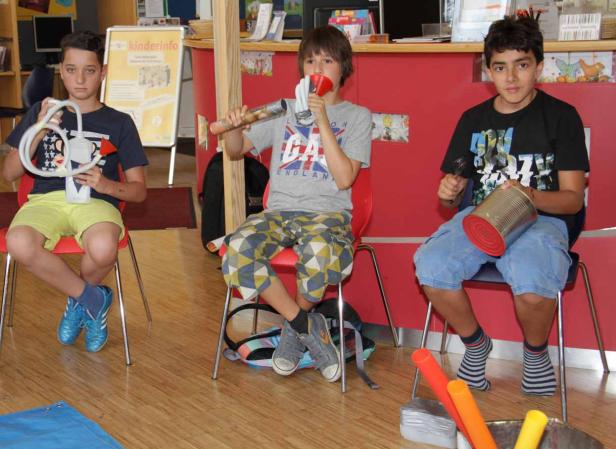 Drei Jungen sitzen auf Stühlen und spielen mit selbstgemachten Musikinstrumenten.