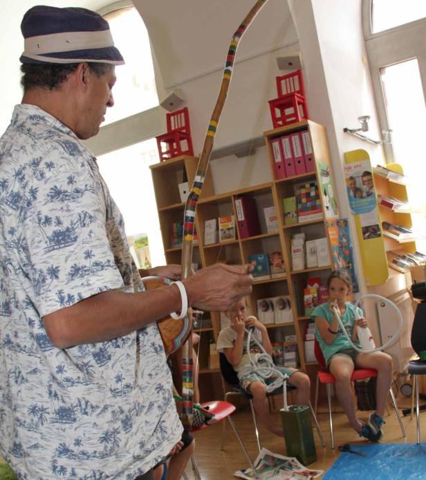 Ein Mann spielt Berimbau vor zwei Kindern in einem Raum.