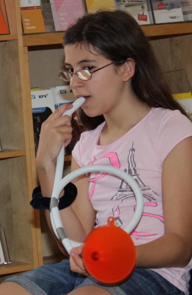 Ein Mädchen mit Brille benutzt ein Atemtrainingsgerät mit einem orangefarbenen Ballon.