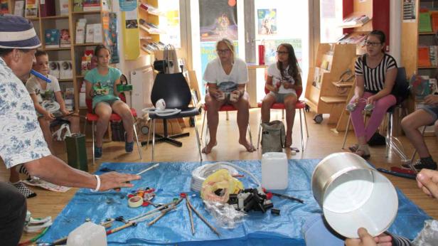 Kinder sitzen um eine blaue Plane mit Bastelmaterialien und werden von einem Mann angeleitet.