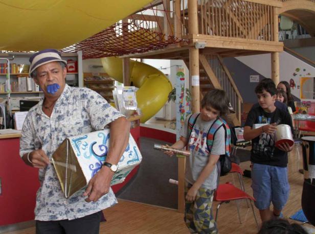 Ein Mann mit einem blauen Mundstück hält eine bemalte Kiste, während Kinder mit Musikinstrumenten im Hintergrund stehen.