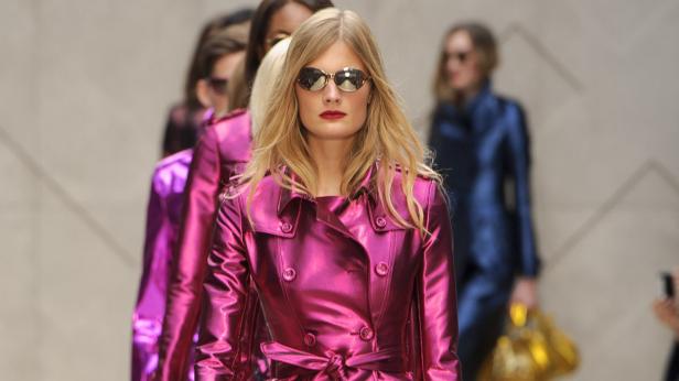 Eine blonde Frau mit Sonnenbrille und pinkfarbenem Mantel läuft auf einem Laufsteg.