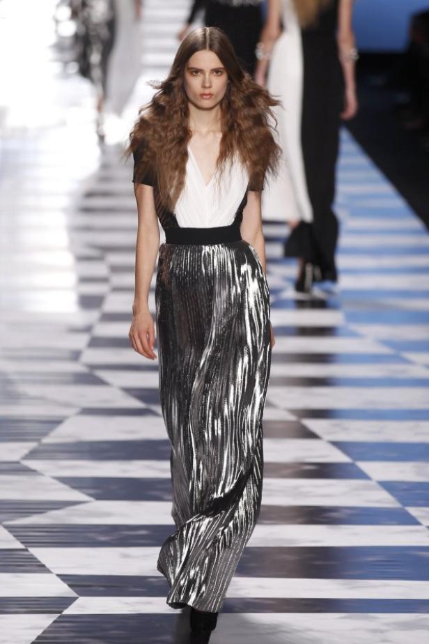 Ein Model präsentiert ein silbernes, plissiertes Abendkleid auf einem Schachbrettmuster-Laufsteg.