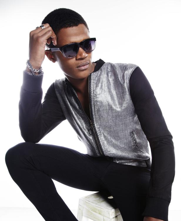 Ein junger Mann mit Sonnenbrille und silberner Jacke posiert auf einem Hocker.