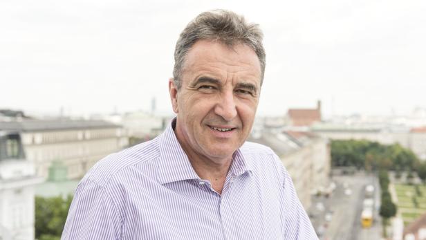 Ein Mann mit einem gestreiften Hemd lächelt vor einem verschwommenen Stadtbild.