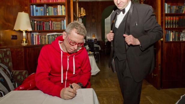 Ein Mann mit roter Jacke schreibt an einem Tisch, während ein Kellner im Hintergrund steht.