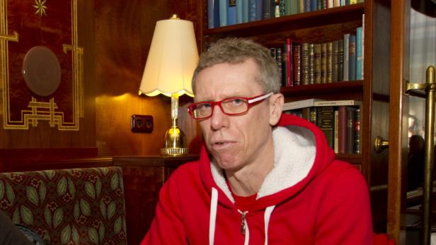 Ein Mann mit roter Brille und rotem Kapuzenpullover sitzt vor einem Bücherregal.