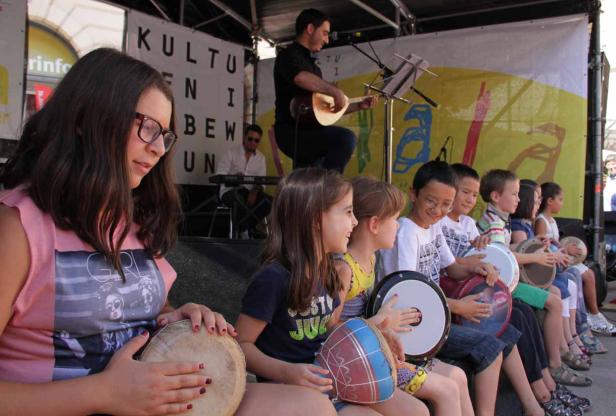 Eine Gruppe Kinder sitzt auf einer Bühne und spielt auf kleinen Trommeln, während ein Mann Gitarre spielt.