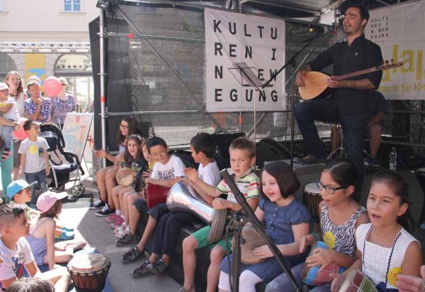 Eine Gruppe Kinder sitzt auf einer Bühne und spielt auf Trommeln, während ein Mann Gitarre spielt.
