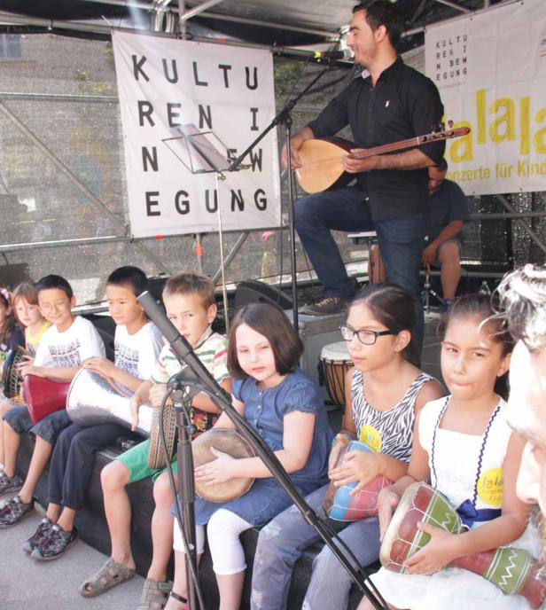 Eine Gruppe Kinder sitzt auf einer Bühne und spielt Trommeln, während ein Mann Saz spielt.