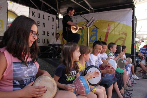 Eine Gruppe Kinder spielt auf einer Bühne mit Trommeln, während ein Mann Gitarre spielt.