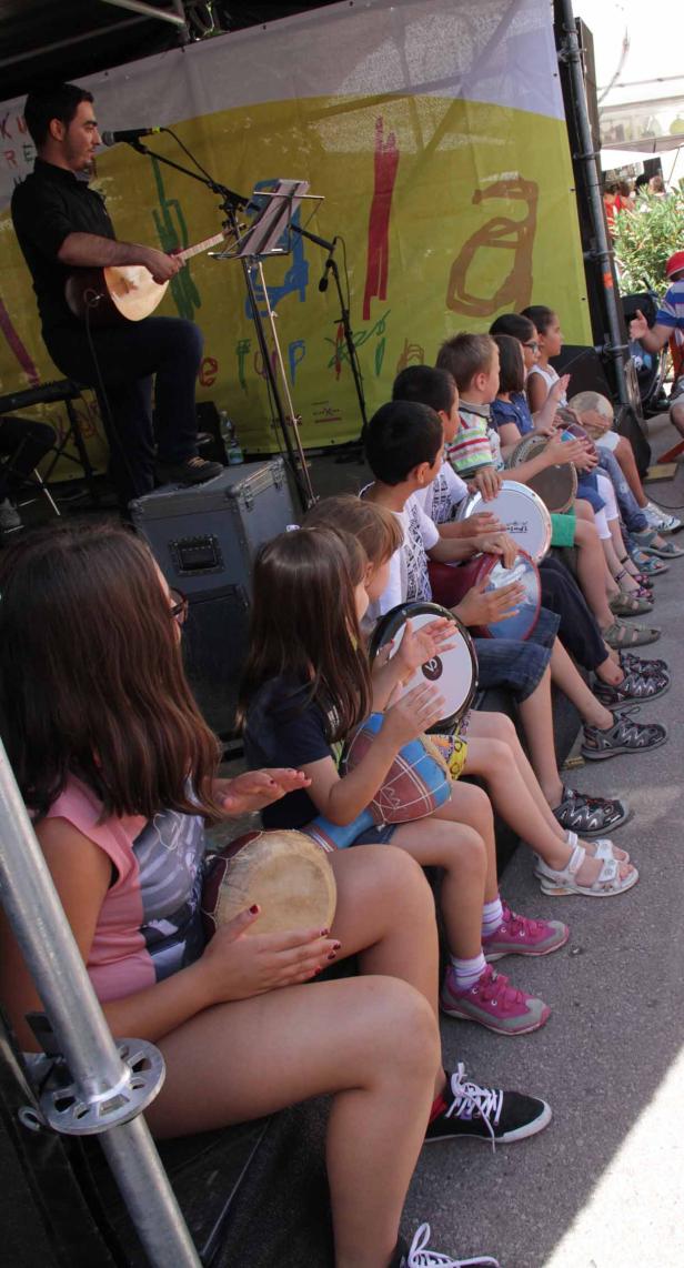 Eine Gruppe Kinder sitzt auf einer Bühne und spielt auf kleinen Trommeln, während ein Mann Gitarre spielt.