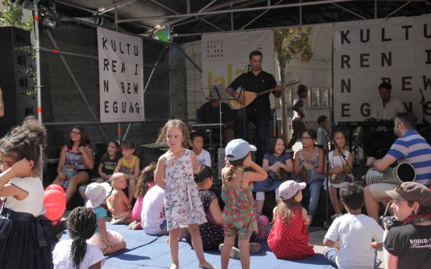 Eine Gruppe von Kindern und Erwachsenen sitzt und musiziert auf einer Bühne vor Bannern.