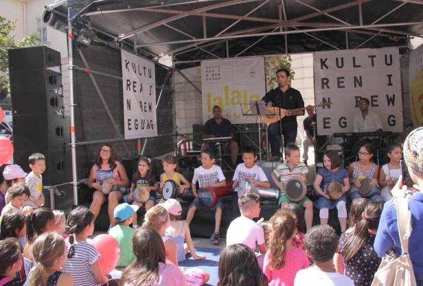 Eine Gruppe von Kindern spielt mit Musikern auf einer Bühne mit Trommeln und Saiteninstrumenten.