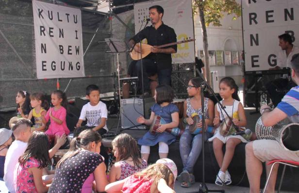 Ein Mann spielt Saz vor einer Gruppe von Kindern mit Trommeln auf einer Bühne.