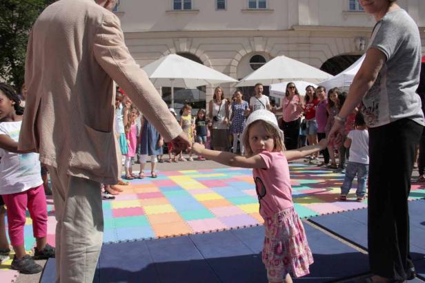 Ein Mädchen hält Händchen mit Erwachsenen auf einer bunten Matte, im Hintergrund weitere Kinder und Erwachsene.