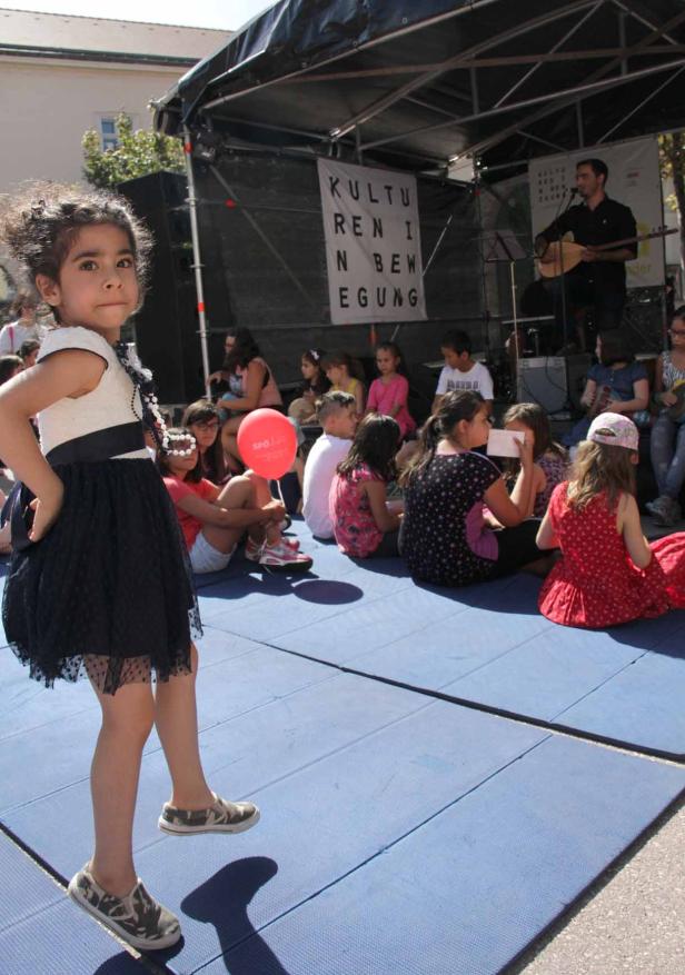 Ein Mädchen steht vor einer Gruppe von Kindern und einem Musiker auf einer Bühne.