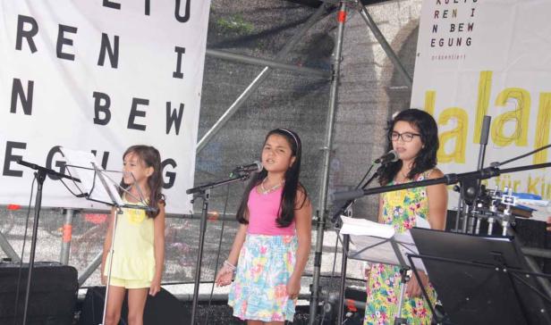 Drei junge Mädchen singen auf einer Bühne vor einem Banner.