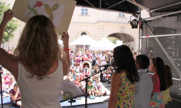 Eine Frau präsentiert eine Kinderzeichnung von einem Kolibri vor einem Publikum auf einer Bühne.